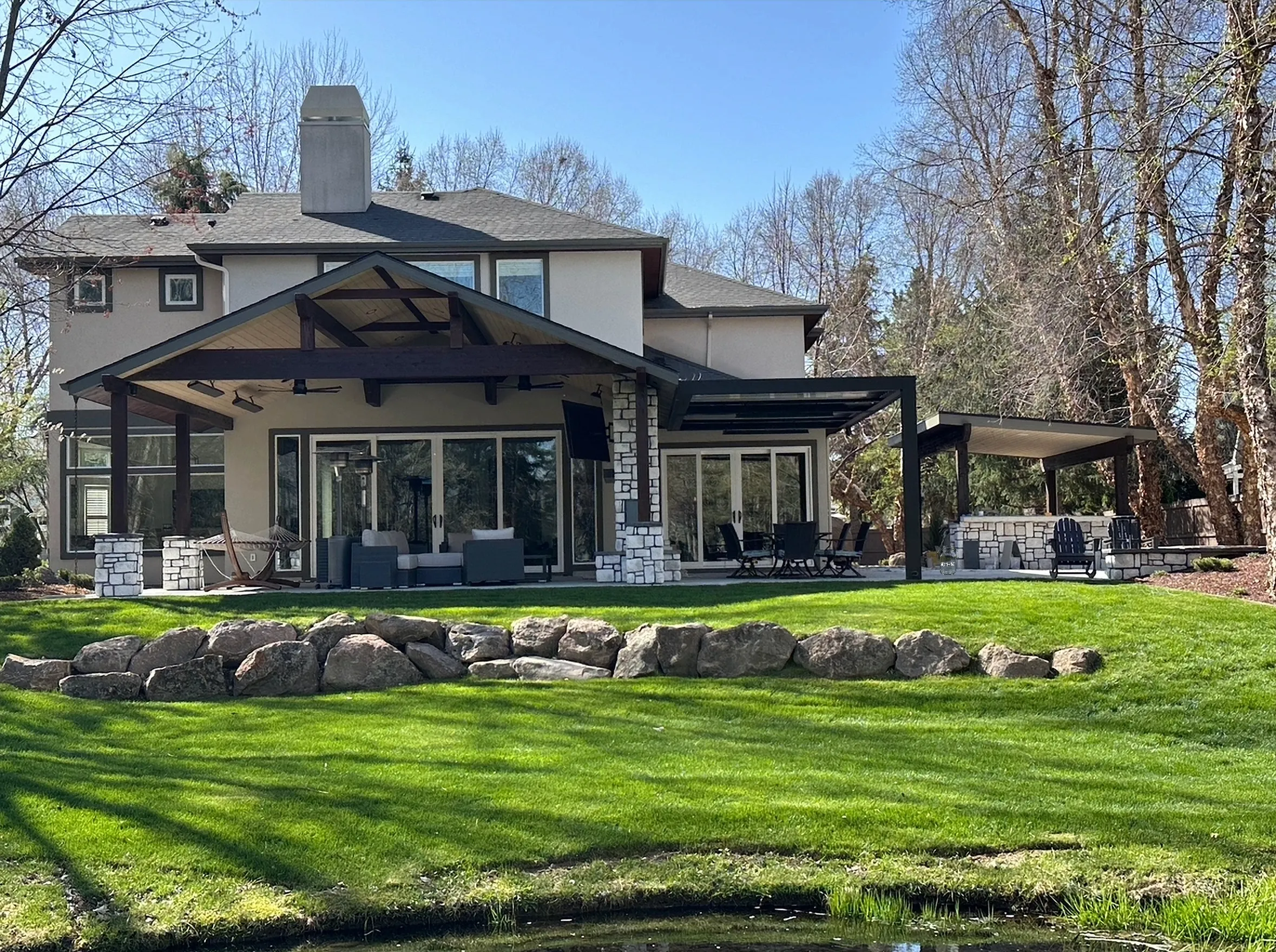 Eagle, Idaho backyard with a timber-frame pavilion, louvered pergola, outdoor kitchen structure, boulder wall and lawn