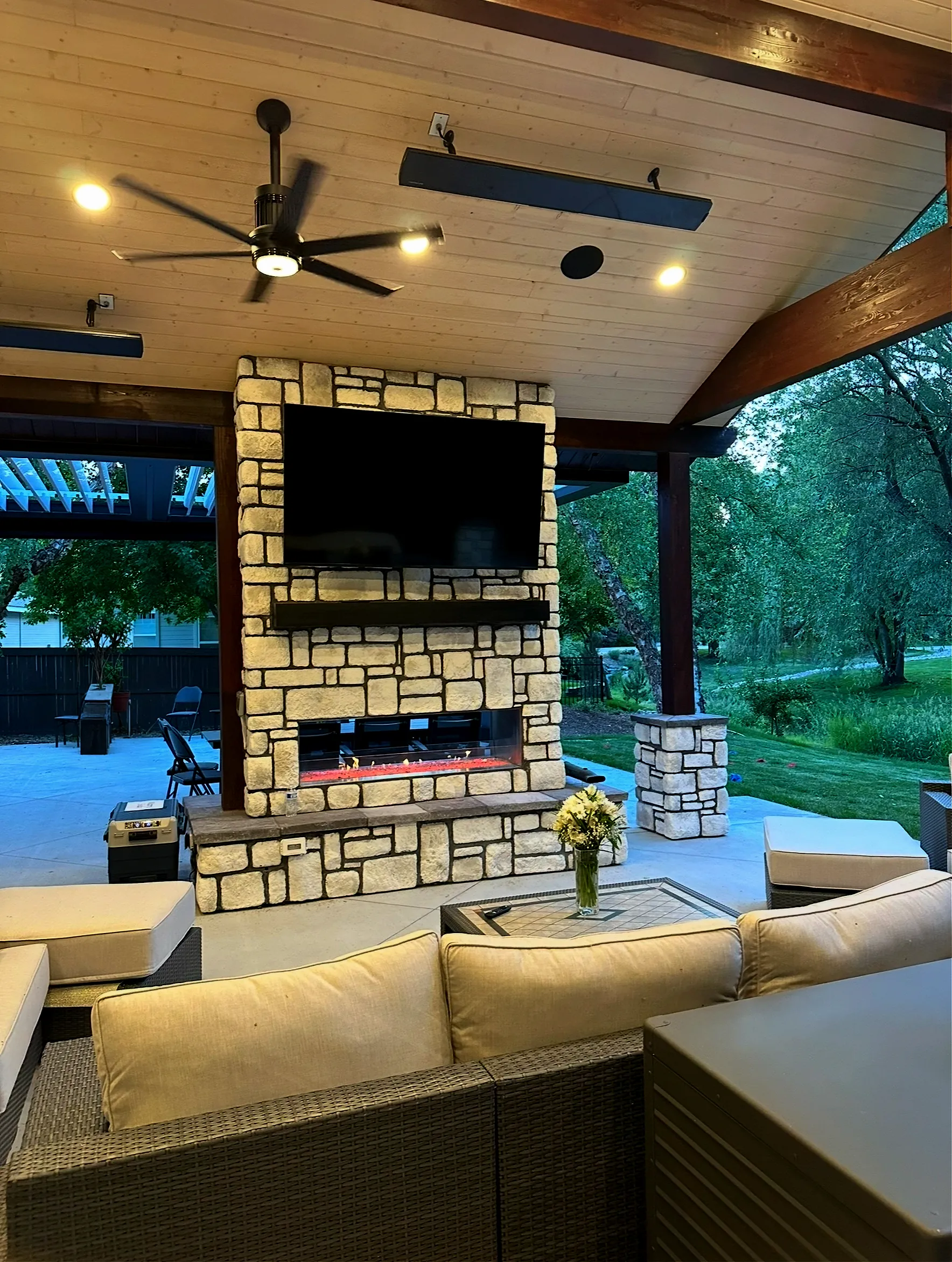 Evening view of a covered patio with a lit stone fireplace, TV and lounge seating