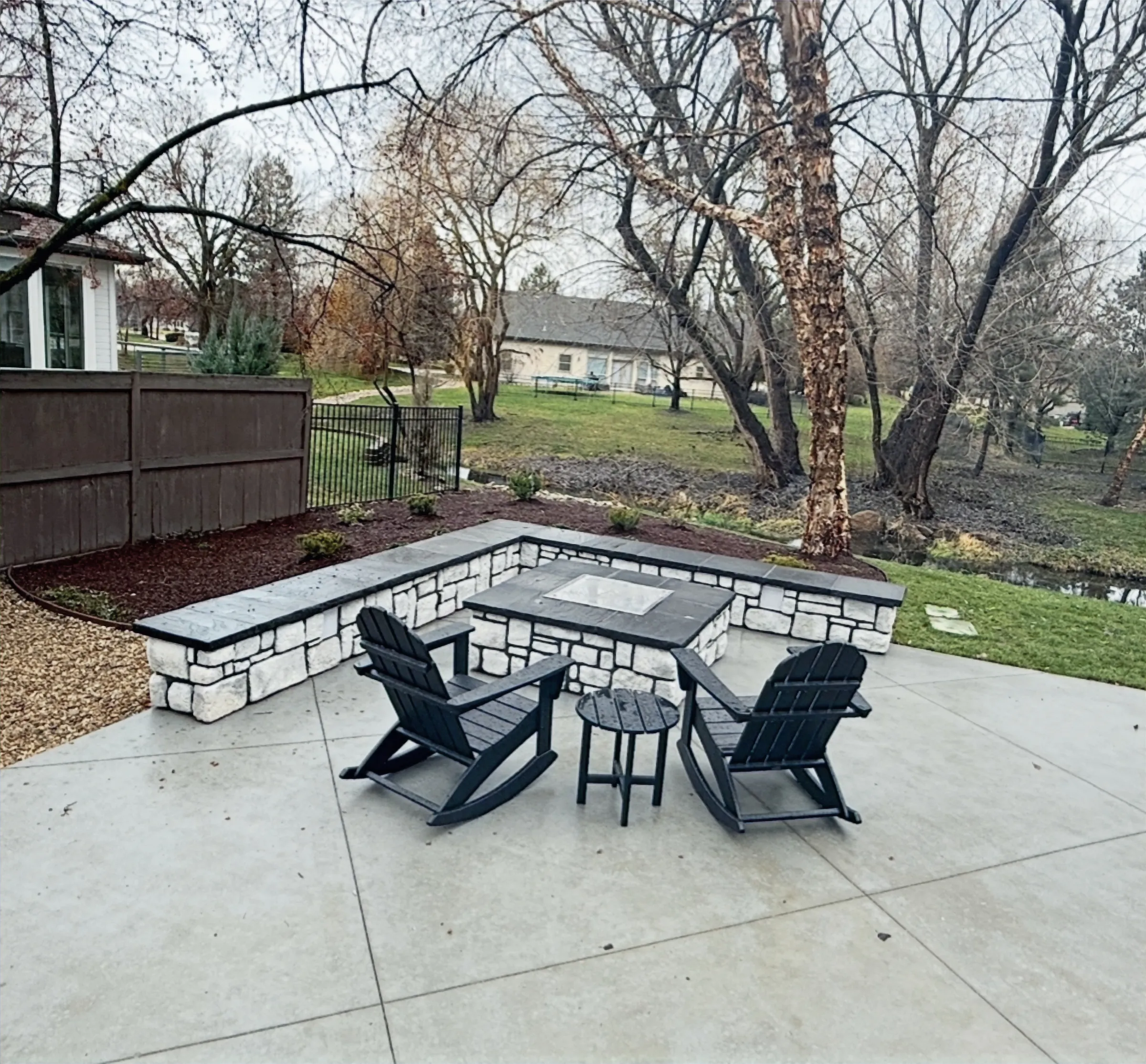 Square fire table inside a stone seat wall on a new concrete patio, with rocking chairs and mature trees